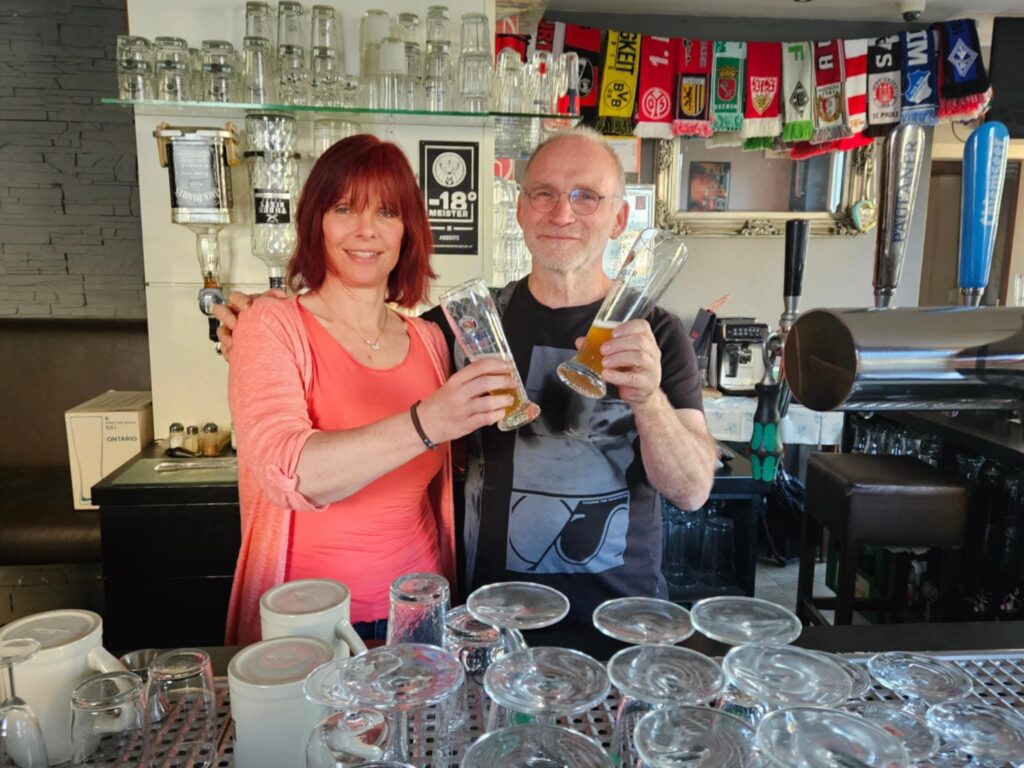 Cerstin und Michael stehen gemeinsam hinterm Tresen der Fußballkneipe "Abseits" in Schriesheim und stoßen mit Biergläsern auf einen schönen gemeinsamen Abend an. Das Bild vermittelt Freundschaft, gute Stimmung und eine entspannte Atmosphäre.