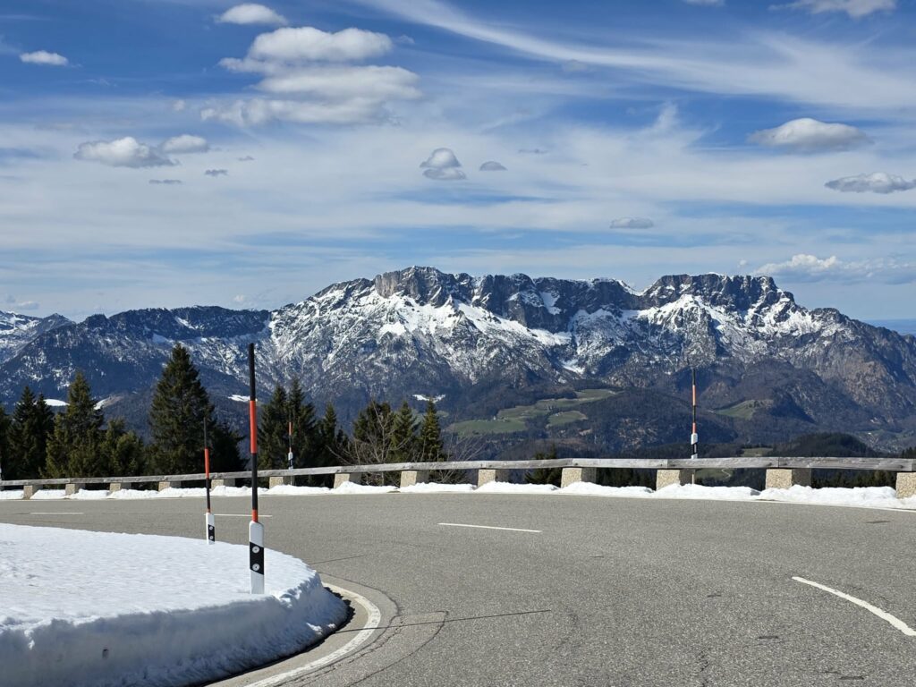 "Panoramablick auf verschneite Berge und die kurvenreiche Rossfeldstraße unter blauem Himmel."